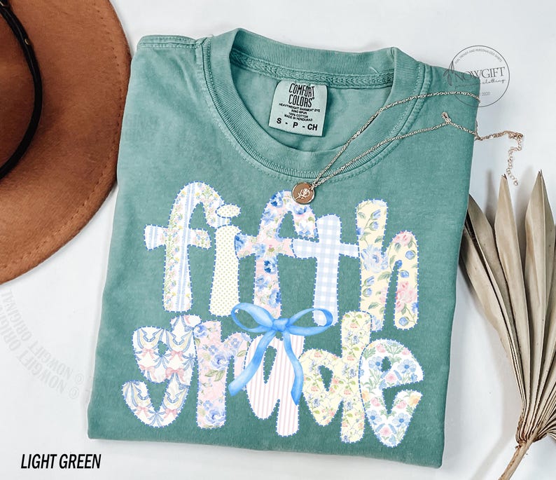 May include: Light green t-shirt with the words "Fifth Grade" in a patchwork design. The letters are decorated with floral and gingham patterns, and a blue bow. A gold necklace is draped across the shirt.