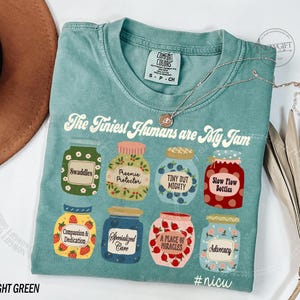 Puede incluir: Camiseta verde claro Comfort Colors con el texto "The Tiniest Humans are My Jam". La camiseta presenta ilustraciones de tarros con varias etiquetas, incluyendo "Swaddles" y "#nicu".