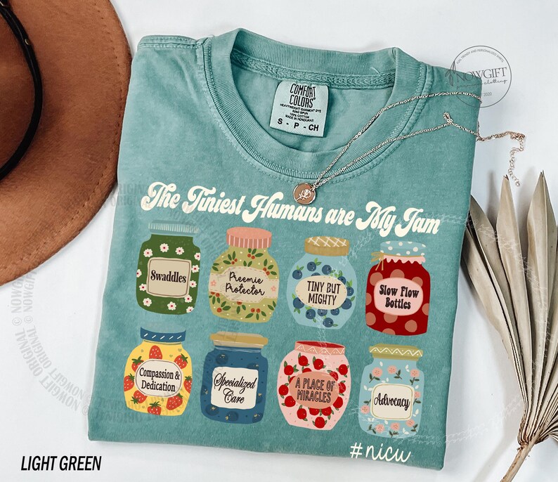 Puede incluir: Camiseta verde claro Comfort Colors con el texto "The Tiniest Humans are My Jam". La camiseta presenta ilustraciones de tarros con varias etiquetas, incluyendo "Swaddles" y "#nicu".