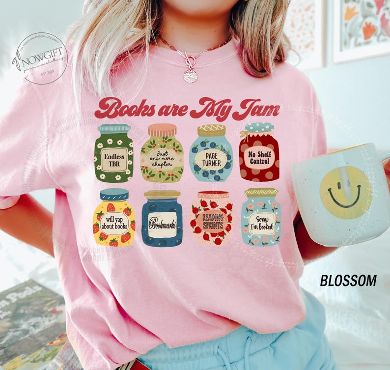 May include: Pale pink sweatshirt featuring the phrase "Books are My Jam" and jar illustrations with book-related text. A mug with a smiley face is held in the hand. The word "BLOSSOM" is printed on the bottom right.
