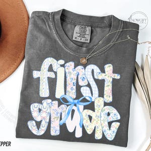 May include: A gray Comfort Colors t-shirt with the words "First Grade" in a patchwork design with floral and gingham patterns. A light blue bow is below the word "First". A gold necklace is on the shirt. The word "Pepper" is at the bottom.
