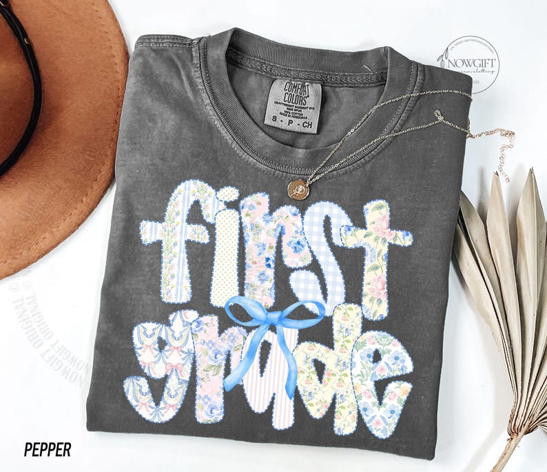 May include: A gray Comfort Colors t-shirt with the words "First Grade" in a patchwork design with floral and gingham patterns. A light blue bow is below the word "First". A gold necklace is on the shirt. The word "Pepper" is at the bottom.
