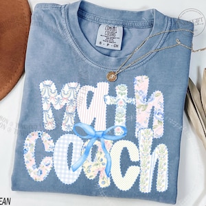 May include: A blue jean Comfort Colors t-shirt with the words "Math Coach" in a patchwork design. The letters are decorated with floral and gingham patterns, and a blue bow is centered below the word "Math". A gold necklace is draped across the shirt.