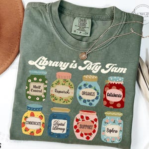 Könnte beinhalten: Ein grünes T-Shirt mit dem Text "Library is My Jam" in einer Retro-Schrift. Das Shirt zeigt eine Illustration von neun Gläsern mit verschiedenen Designs und Texten, darunter "Shelf Control", "Research", "Organize", "Collaborate", "Communicate", "Digital Literacy", "Support" und "Explore".