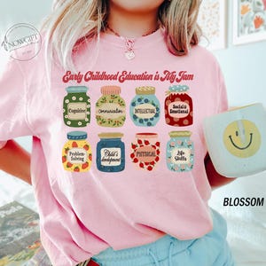 Puede incluir: Camiseta rosa con el texto "Early Childhood Education is My Jam" y un gr&aacute;fico de tarros con diferentes aspectos de la educaci&oacute;n de la primera infancia, como la cognici&oacute;n, la comunicaci&oacute;n, la intelectual, la social y la emocional, la resoluci&oacute;n de problemas, el desarrollo del ni&ntilde;o, la f&iacute;sica y las habilidades para la vida.