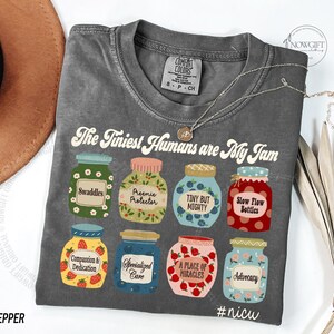 Puede incluir: Una camiseta gris con el texto "The Tiniest Humans are My Jam" e ilustraciones de tarros de mermelada. Los tarros tienen etiquetas como "Swaddles" y "Advocacy". La camiseta est&aacute; etiquetada como "Pepper".