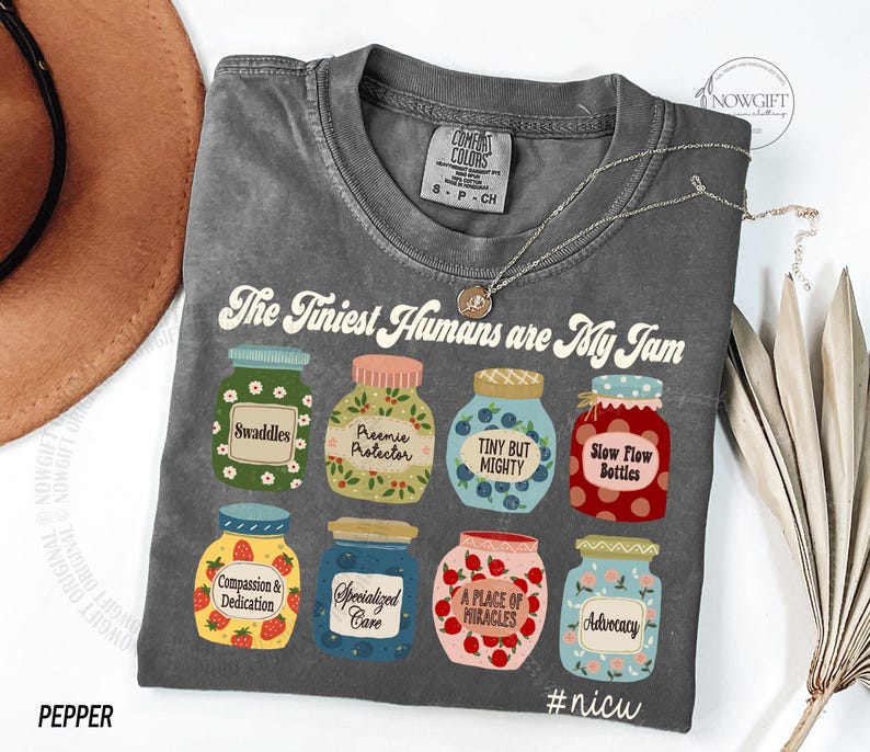 Puede incluir: Una camiseta gris con el texto "The Tiniest Humans are My Jam" e ilustraciones de tarros de mermelada. Los tarros tienen etiquetas como "Swaddles" y "Advocacy". La camiseta est&aacute; etiquetada como "Pepper".