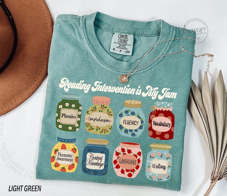 May include: Light green t-shirt with the text "Reading Intervention is My Jam" and illustrations of jars with different reading skills, such as "phonics", "comprehension", "fluency", and "vocabulary".