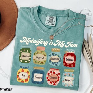 May include: Light green t-shirt with the text "Midwifery is My Jam" and illustrations of jars labeled with words like "Education" and "Joy." A silver necklace with a pendant is draped across the shirt.