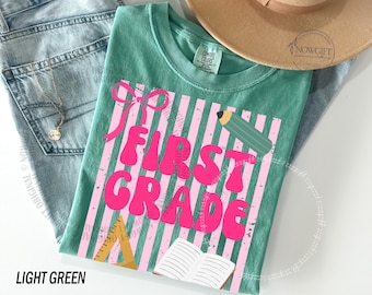 Op de afbeelding: Lichtgroen T-shirt met de woorden "FIRST GRADE" in felroze, tegen een roze en wit gestreepte achtergrond. Een strik, potlood, liniaal en open boek zijn ook te zien. Het shirt wordt getoond met een blauwe jeans en een bruine hoed.
