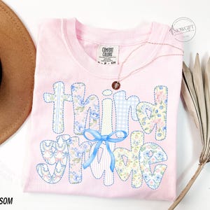 Puede incluir: Una camiseta rosa claro Comfort Colors con las palabras "Think Spring" en un dise&ntilde;o de patchwork. Las letras est&aacute;n decoradas con estampados florales, cuadros vichy y un lazo azul. Tambi&eacute;n se ven un collar dorado y un sombrero marr&oacute;n.