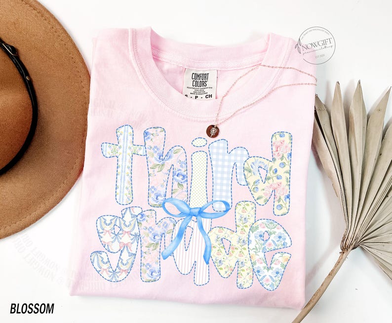 Puede incluir: Una camiseta rosa claro Comfort Colors con las palabras "Think Spring" en un dise&ntilde;o de patchwork. Las letras est&aacute;n decoradas con estampados florales, cuadros vichy y un lazo azul. Tambi&eacute;n se ven un collar dorado y un sombrero marr&oacute;n.