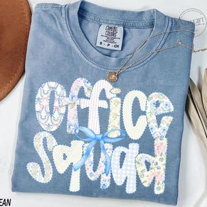 May include: A blue jean Comfort Colors t-shirt with the words "Office Squad" in a patchwork design. The letters are made of floral and patterned fabrics. A blue bow is centered below the word "Office". A gold necklace with a pendant rests on the shirt.