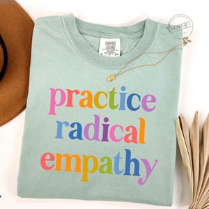 May include: A light green t-shirt with the words "practice radical empathy" in colorful letters. A gold necklace and a brown hat are next to the shirt. The shirt is made of a soft material.