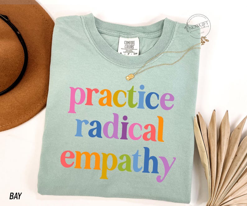 May include: A light green t-shirt with the words "practice radical empathy" in colorful letters. A gold necklace and a brown hat are next to the shirt. The shirt is made of a soft material.