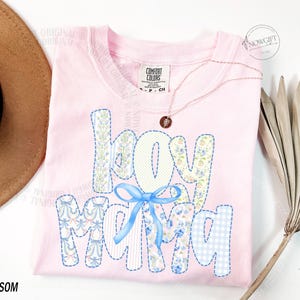 Puede incluir: Camiseta rosa claro con la frase "Hey Mama" en un dise&ntilde;o de patchwork. Las letras est&aacute;n decoradas con estampados florales y un lazo azul. Tambi&eacute;n se ven un collar dorado y un sombrero marr&oacute;n.