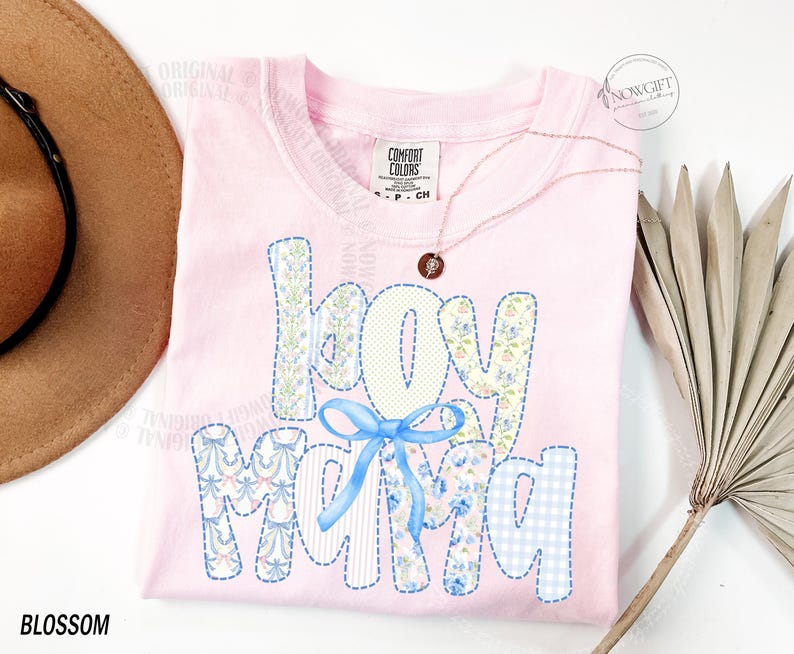 Puede incluir: Camiseta rosa claro con la frase "Hey Mama" en un dise&ntilde;o de patchwork. Las letras est&aacute;n decoradas con estampados florales y un lazo azul. Tambi&eacute;n se ven un collar dorado y un sombrero marr&oacute;n.