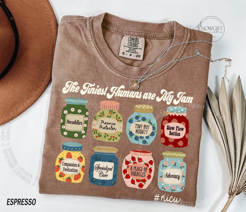 Puede incluir: Camiseta marr&oacute;n con la frase "The Tiniest Humans are My Jam" e ilustraciones de tarros. Los tarros tienen etiquetas como "Swaddles" y "Advocacy". Tambi&eacute;n se ven un collar plateado y un sombrero marr&oacute;n.