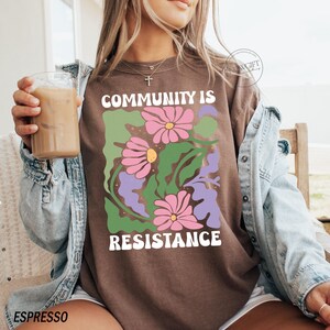 May include: A brown t-shirt with a graphic design of flowers and leaves. The text "Community is Resistance" is printed on the shirt.
