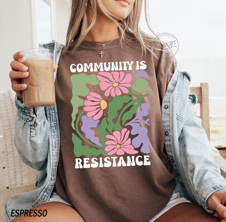 May include: A brown t-shirt with a graphic design of flowers and leaves. The text "Community is Resistance" is printed on the shirt.