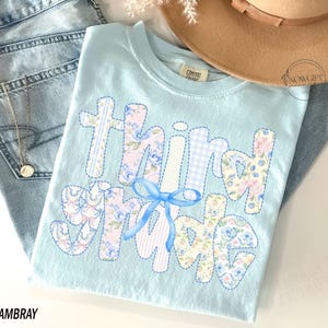 Puede incluir: Una camiseta azul claro con las palabras "Third Grade" en un dise&ntilde;o de patchwork. Las letras est&aacute;n decoradas con estampados florales y de cuadros, y un lazo azul. La camiseta se muestra con unos vaqueros azules y un sombrero marr&oacute;n.