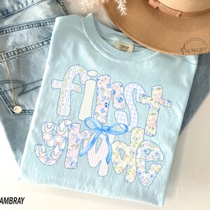 May include: A light blue t-shirt with the words "first grade" in a patchwork design. The letters are made of floral, striped, and dotted fabrics. A blue bow is at the bottom. The shirt is displayed with light blue jeans and a tan hat.