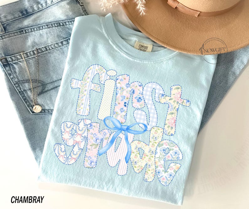 May include: A light blue t-shirt with the words "first grade" in a patchwork design. The letters are made of floral, striped, and dotted fabrics. A blue bow is at the bottom. The shirt is displayed with light blue jeans and a tan hat.
