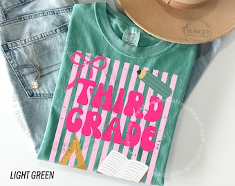Op de afbeelding: Lichtgroen t-shirt met de roze woorden "THIRD GRADE", tegen een roze en wit gestreepte achtergrond. Het shirt heeft een strik, potlood, liniaal en open boek afbeelding. Het shirt wordt getoond met een spijkerbroek en een bruine hoed.