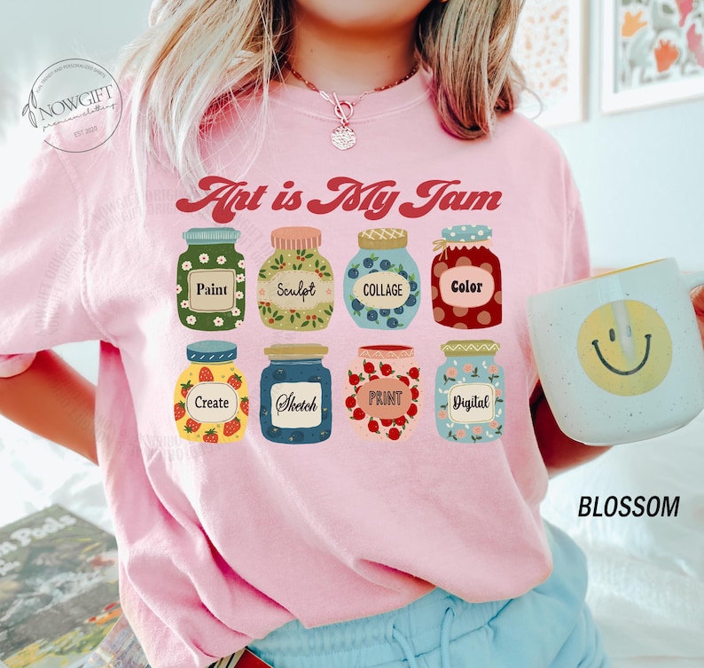 Op de afbeelding: Roze T-shirt met de tekst "Art is My Jam" en een afbeelding van acht potten met verschillende kunstgerelateerde woorden erop, zoals "Verven", "Beeldhouwen", "Collage", "Kleur", "Cre&euml;ren", "Schetsen", "Drukken" en "Digitaal".