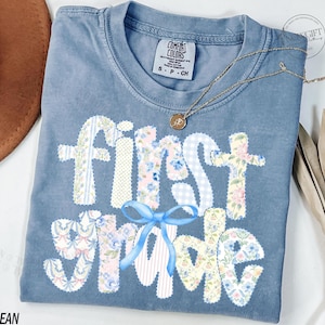 May include: Blue jean Comfort Colors t-shirt with "First Grade" in patchwork letters. The letters are decorated with floral and gingham patterns, and a blue bow. A gold necklace with a pendant is on the shirt.