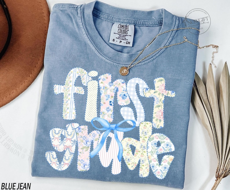 May include: Blue jean Comfort Colors t-shirt with "First Grade" in patchwork letters. The letters are decorated with floral and gingham patterns, and a blue bow. A gold necklace with a pendant is on the shirt.