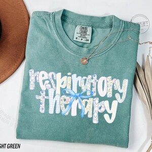 Pu&ograve; includere: T-shirt verde chiaro con la scritta "respiratory therapy" in un design patchwork con un fiocco azzurro. Una collana d'oro con un ciondolo &egrave; drappeggiata sulla maglietta. La maglietta &egrave; piegata su una superficie bianca.