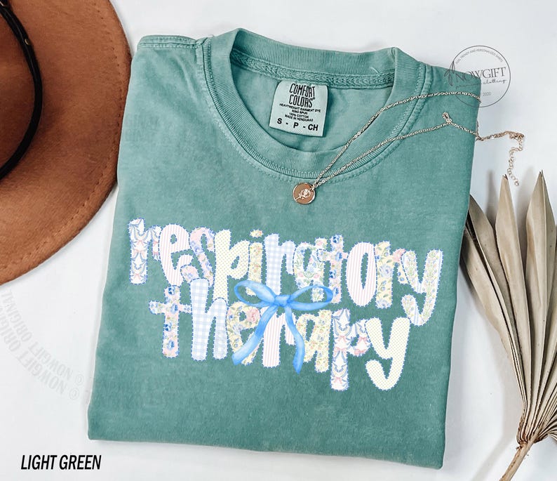 Pu&ograve; includere: T-shirt verde chiaro con la scritta "respiratory therapy" in un design patchwork con un fiocco azzurro. Una collana d'oro con un ciondolo &egrave; drappeggiata sulla maglietta. La maglietta &egrave; piegata su una superficie bianca.