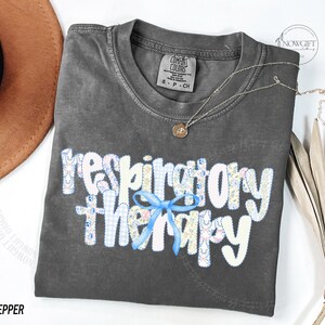 Pu&ograve; includere: Una maglietta grigio scuro con le parole "respiratory therapy" in un design patchwork e un fiocco blu. Una collana d'oro con un ciondolo &egrave; sulla maglietta. La maglietta &egrave; etichettata "Pepper".