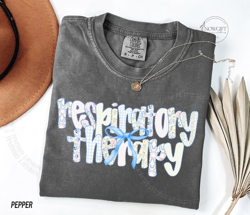 Pu&ograve; includere: Una maglietta grigio scuro con le parole "respiratory therapy" in un design patchwork e un fiocco blu. Una collana d'oro con un ciondolo &egrave; sulla maglietta. La maglietta &egrave; etichettata "Pepper".