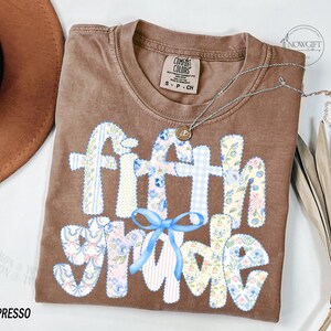 May include: Espresso-colored t-shirt with the words "Fifth Grade" in a patchwork design. The letters are made of floral and gingham patterns, with a light blue bow. A silver necklace with a pendant is draped across the shirt.