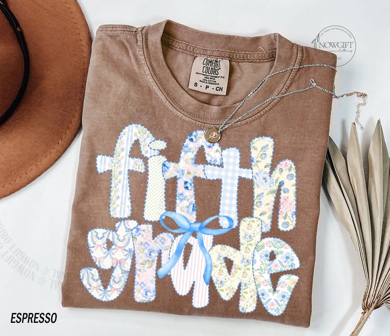 May include: Espresso-colored t-shirt with the words "Fifth Grade" in a patchwork design. The letters are made of floral and gingham patterns, with a light blue bow. A silver necklace with a pendant is draped across the shirt.