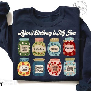 May include: Navy blue sweatshirt with a graphic of jars of jam, each labeled with a stage of labor and delivery. The text "Labor & Delivery is My Jam" is printed above the jars.