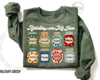 Op de afbeelding: Militairgroen sweatshirt met de tekst "Activities are My Jam" en illustraties van acht jampotten, elk gelabeld met een positief woord zoals "Support" en "Creative".