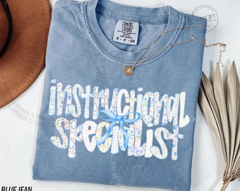 Op de afbeelding: Blauwe Comfort Colors-t-shirt met de woorden "Instructional Specialist" in een bloemenpatroon. Een lichtblauwe strik is gecentreerd onder de tekst. Een gouden ketting met een hanger is over het shirt gedrapeerd.