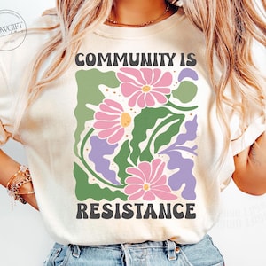 May include: A white t-shirt with a graphic design of pink and green flowers and the text "Community is Resistance".