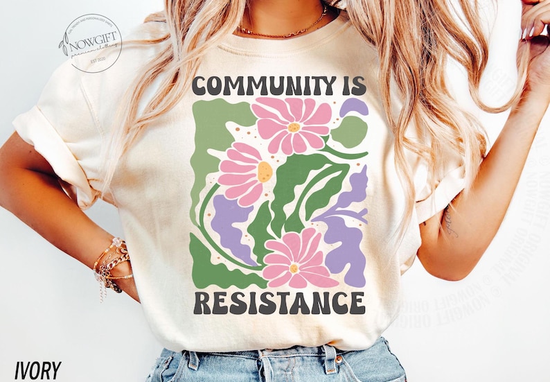 May include: A white t-shirt with a graphic design of pink and green flowers and the text "Community is Resistance".