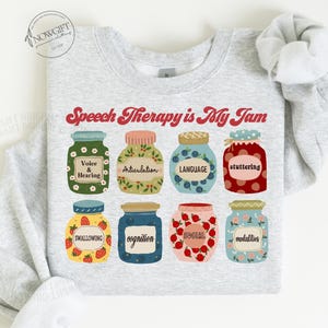 May include: A light gray sweatshirt with the text "Speech Therapy is My Jam" printed in red. The text is surrounded by illustrations of jars with different speech therapy related terms, such as "Voice & Hearing", "Articulation", "Language", "Stuttering", "Swallowing", "Cognition", "Social", and "Modalities".