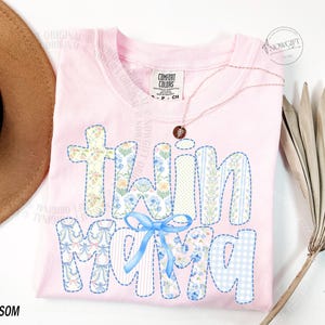 Puede incluir: Una camiseta rosa claro con las palabras "Twin Mom" en un dise&ntilde;o de patchwork. Las letras presentan patrones florales, a rayas y de cuadros con un lazo azul. La camiseta tiene un cuello redondo y una peque&ntilde;a etiqueta que dice "Comfort Colors".
