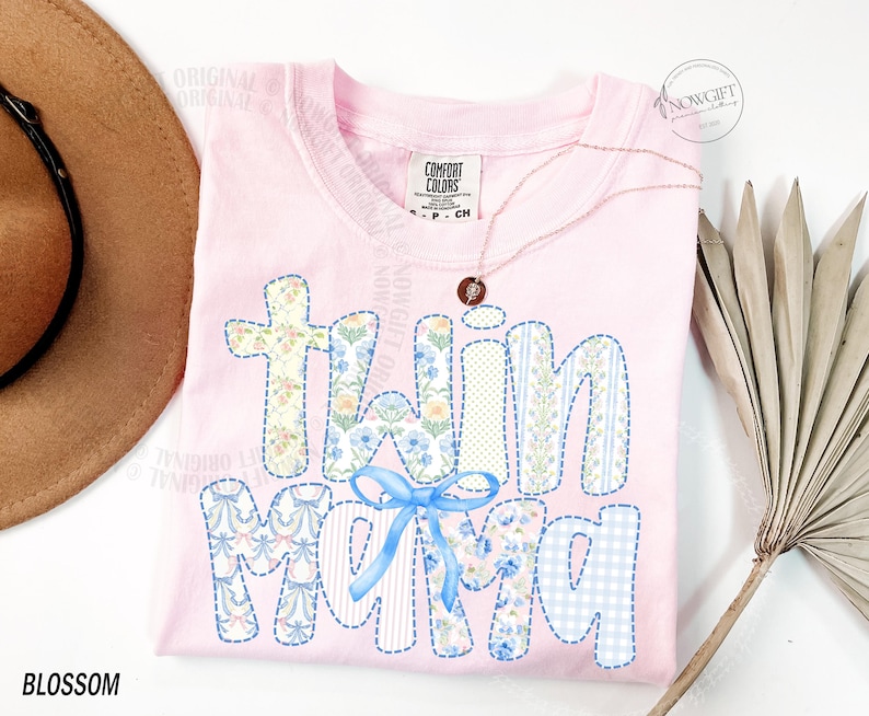 Puede incluir: Una camiseta rosa claro con las palabras "Twin Mom" en un dise&ntilde;o de patchwork. Las letras presentan patrones florales, a rayas y de cuadros con un lazo azul. La camiseta tiene un cuello redondo y una peque&ntilde;a etiqueta que dice "Comfort Colors".
