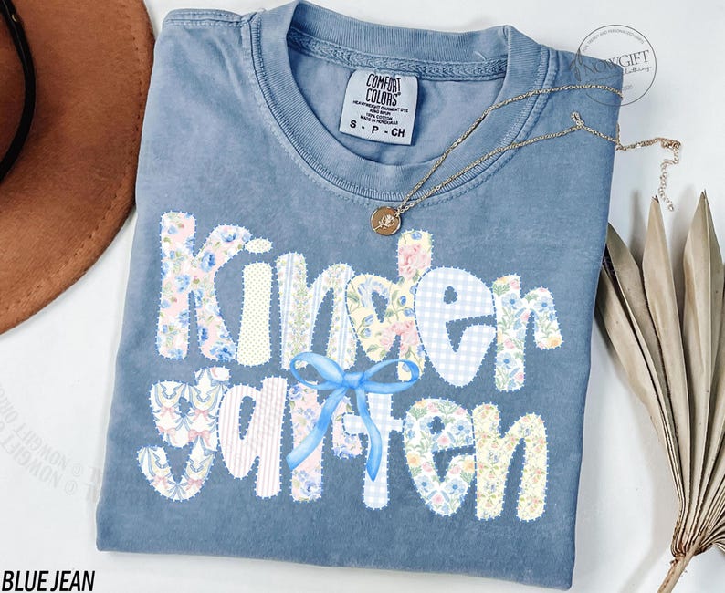 Pu&ograve; includere: T-shirt blu jeans Comfort Colors con "Kindergarten" in lettere patchwork. Le lettere sono decorate con motivi floreali e gingham, e un fiocco blu. Una collana d'oro con un ciondolo &egrave; drappeggiata sulla maglietta.