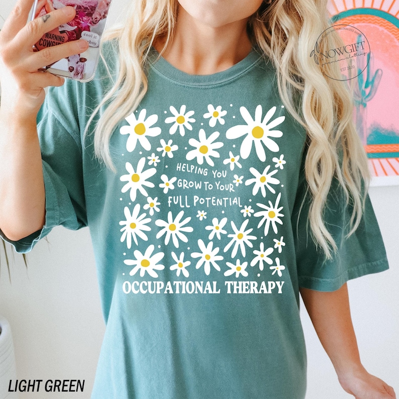 May include: Light green t-shirt featuring a daisy design with the text "Helping You Grow To Your Full Potential" and "Occupational Therapy" in white. The shirt is a comfortable, casual style.
