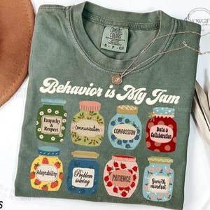 Peut inclure: Un t-shirt vert avec le texte "Behavior is My Jam" dans une police rétro. Le t-shirt présente un motif de bocaux avec différents mots liés à un comportement positif, tels que "Empathie et Respect", "Communication", "Compassion", "Données et Collaboration", "Adaptabilité", "Résolution de problèmes", "Patience" et "Mentalité de croissance".