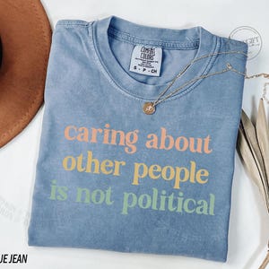 May include: A blue jean t-shirt with the text "caring about other people is not political" printed in rainbow colors.