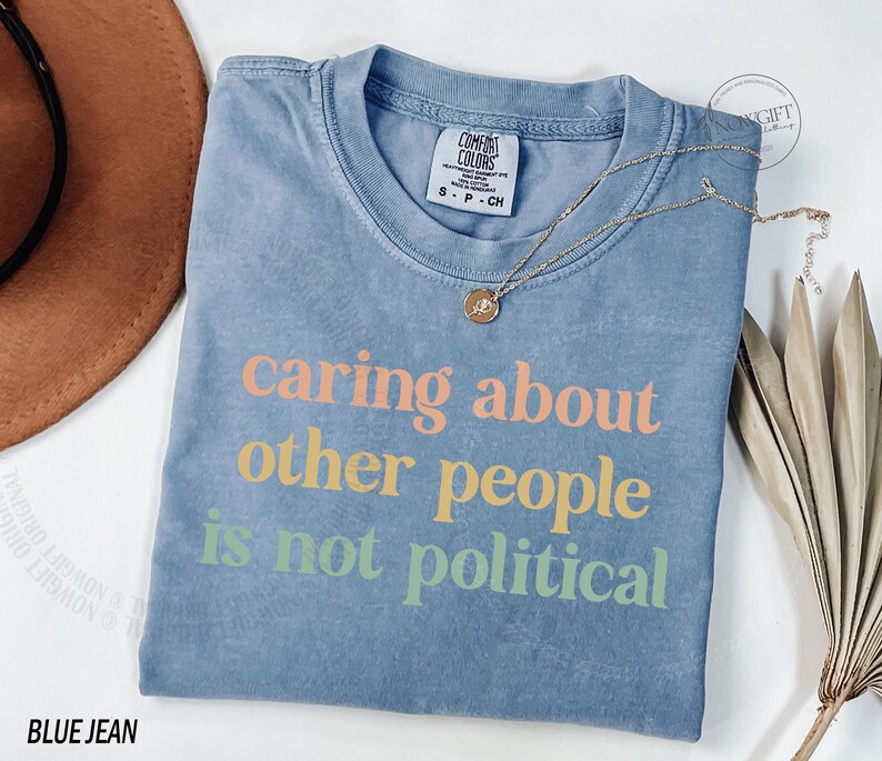 May include: A blue jean t-shirt with the text "caring about other people is not political" printed in rainbow colors.